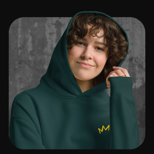Lächelnde Person mit lockigen Haaren trägt einen dunkelgrünen Hoodie mit gelber Krone auf der linken Brust. Hintergrund in grauer Betonoptik.
