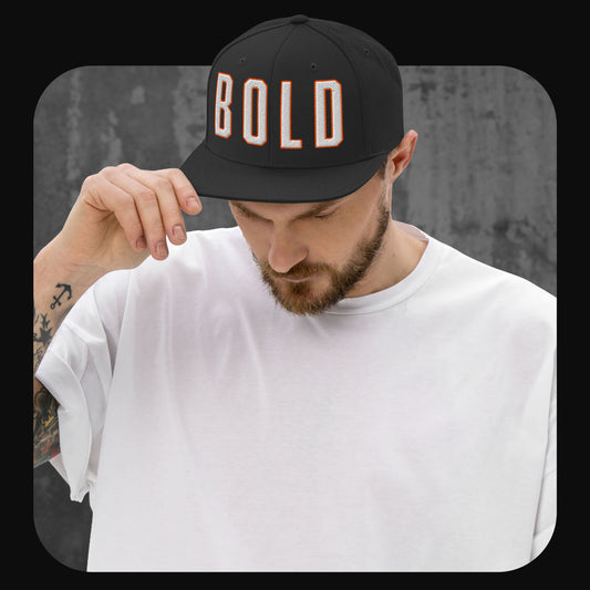 Mann mit schwarzer Snapback-Cap mit dem Wort „BOLD“ in weißen Blockbuchstaben mit oranger Umrandung. Kombiniert mit weißem T-Shirt, Person schaut nach unten und fasst den Schirm der Cap an.