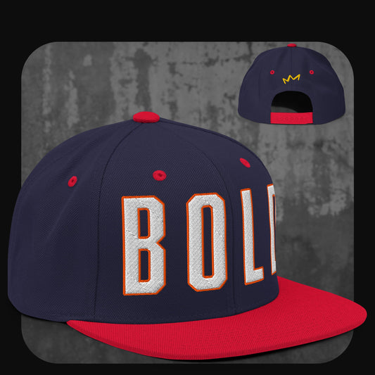 Dunkelblaue Snapback-Cap mit roter Krempe, roten Ösen und rotem Knopf. Vorne gestickter „BOLD“-Schriftzug in Weiß mit oranger Umrandung, hinten gelbes Kronenlogo.
