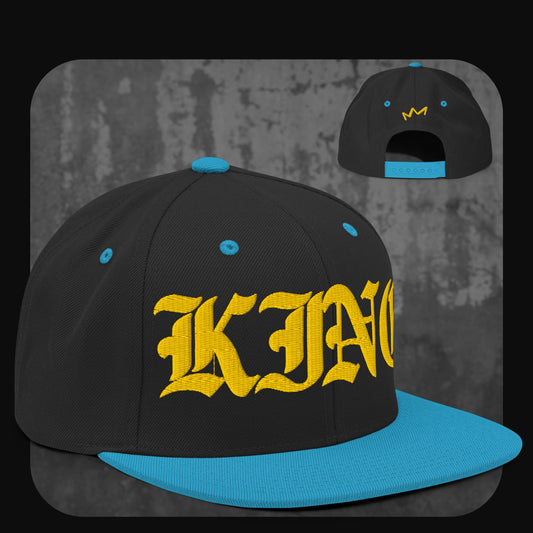 Schwarze Snapback-Cap mit blauer Krempe, blauen Ösen und blauem Knopf. Auf der Vorderseite steht „KING“ in gelber Frakturschrift, auf der Rückseite ist eine kleine gelbe Krone gestickt.
