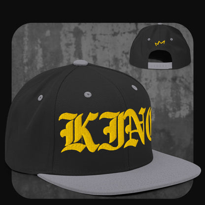 Schwarze Snapback-Cap mit grauer Krempe, grauen Ösen und grauem Knopf. Auf der Vorderseite gelber „KING“-Schriftzug, auf der Rückseite kleine gelbe Krone.