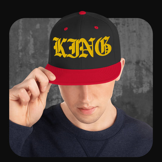 Person trägt eine schwarze Snapback-Cap mit roter Krempe, roten Ösen und rotem Knopf. Auf der Vorderseite steht in großer gelber Frakturschrift das Wort „KING“.