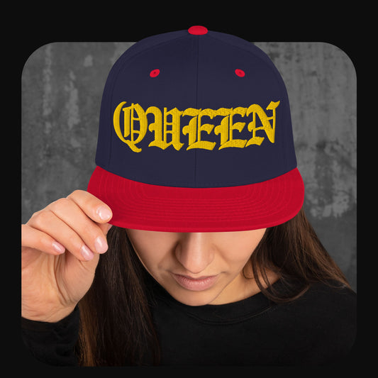 Frau trägt eine dunkelblaue Snapback-Cap mit roter Krempe, roten Ösen und rotem Knopf. Auf der Vorderseite steht in großer gelber Frakturschrift das Wort „QUEEN“.