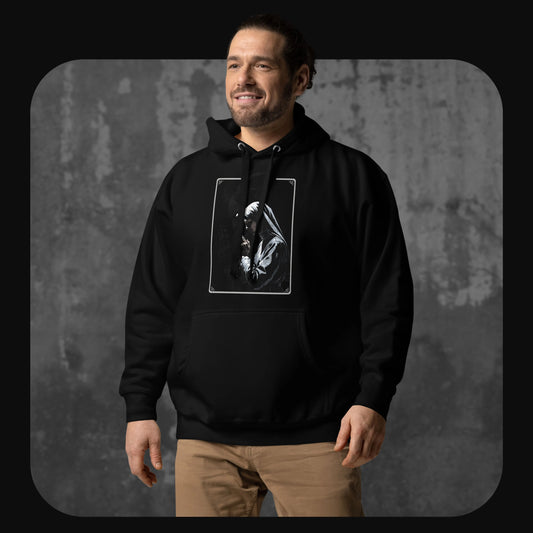 Smiling man wearing a black hoodie featuring a grayscale illustration of a praying nun on the front, standing against a concrete backdrop.